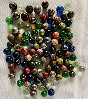 Top-down view of approximately 100 assorted colored glass marbles arranged on a white textured surface showing various colors and speckled patterns.