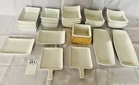 Top view showing full lot of white ceramic and porcelain dinnerware pieces, including multiple small and medium square bowls and dishes, rectangular plates, two handled serving dishes and a dipping bowl in a wicker holder.
