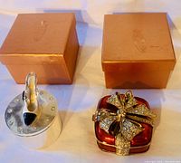 Two trinket boxes with tan presentation boxes behind them. Left box is round silver-tone with shoe figurine on top. Right box is red enamel with jewelled bow.