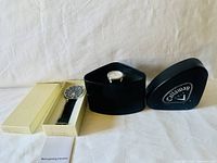 Two men's watches displayed with their cases and packaging on light background. One black-faced watch in cream box with instruction sheet. Other with white face in Callaway black case.