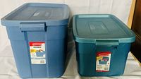 Front view of two blue Rubbermaid Roughneck storage totes with lids, showing labels and size difference.