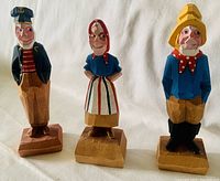 Three hand-carved wooden figurines of elderly characters in colorful clothing, standing on square wooden bases, signed Hannah.