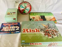 Photo of four boxed classic board games Risk, Backgammon, Rummikub, and a Holiday Wheel of Kindness spinner, arranged on a white surface.