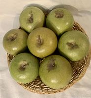 7 artificial green apple fruit arranged in a wicker tray viewed from above showing realistic texture and stems.