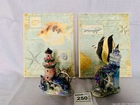 Overall view of 2 ceramic lighthouse figurines with dolphins in front of two tropical fish prints, showing size and condition.