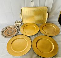Photo showing four gold charger plates, one gold rectangular tray with handles, one decorative round plate, and small set of metal clips or hooks to the left.