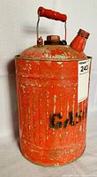 Front side of vintage red metal gas can showing wooden handle and worn red paint with 'GASOLINE' stenciled in black partially visible.