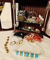 Open jewelry box showing various bracelets, rings, earrings, necklaces, brooch inside on glass shelves and surface