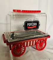 Front angle view of red wagon popcorn maker showing clear plastic lid, red trim lid handle, and wagon wheels.