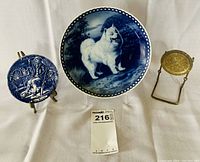 Photo showing two blue and white porcelain collectible plates and a metal holder stand. Left plate (4 inch) is Wedgwood Etruria England 'The Games Keeper' plate featuring an outdoor scene with a seated man. Center plate (7.5 inch) shows white dog artwork by Tove Svendsen for Kunsthandwerk.