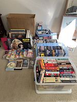 Overall lot of vinyl albums stacked, 8-track tapes, cassette tapes, and VHS tapes in tubs and a cardboard box on carpet.
