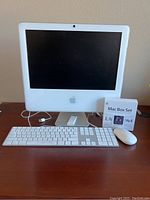 White Apple iMac with keyboard, mouse, remote, and Mac Box Set software on a wooden surface.
