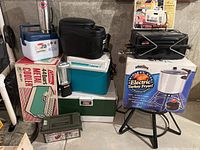 Overview of camping and outdoor items showing stack of Coleman coolers including metal and plastic variations, black portable propane grill, and electric turkey fryer on metal stand