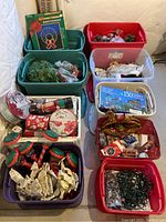 Nine plastic tubs filled with assorted Christmas decoration items including garlands, tins, ribbon bows, ornaments, string lights, and holiday-themed tins.
