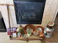 Wide view of all items placed in front of fireplace including wooden tool, brass pans, tins, metallic and glassware decor, small plant and framed notes.