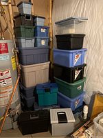 Stack of plastic tubs of various sizes and colors including grey, blue, beige, and black with some having metal latches and others smooth lids. Lot contains approximately 23 used but clean plastic tubs.