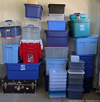 Stack of assorted empty plastic storage tubs in various colors alongside a black metal-trimmed storage trunk on the floor.