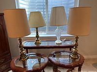 Four table lamps on wooden tables near window; three brass with beige shades and one etched glass with white shade