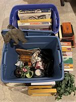 Two large plastic tubs: one filled with several boxed board games, including Wheel of Fortune, Parcheesi, Sorry, Yahtzee; other contains various decor items such as elephant figurine, floral pieces, and small household objects.