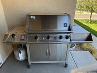 Front view of Members Mark stainless steel 4-burner gas grill showing control knobs, propane tank, and side shelves.