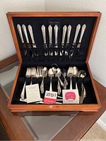 Wooden case open displaying multiple pieces of silver plated flatware including knives, forks, spoons, and serving pieces arranged in black velvet-like interior with elastic straps.