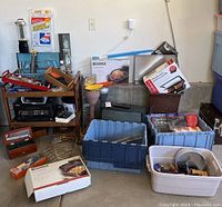 Wide view of large lot with vintage bar cart, plastic tubs, boxed kitchen appliances, outdoor floor mat, printer, and other household items on concrete floor against wall.