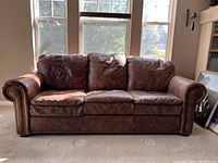 Front view of brown leather sofa placed in front of large windows showing overall shape, three seat cushions, three back cushions, rounded arms with brass studs.