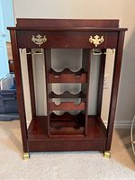 Front view of wood beverage cart showing drawer with brass handles, removable tray on top, three bottle holders, and metal holders on sides.