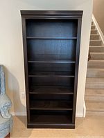 Front view of tall dark brown wood bookcase with five shelves and decorative molding on top, showing minor surface wear.