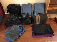 Overview of all backpacks and bags arranged on the floor against wall, showing variety of brands and types