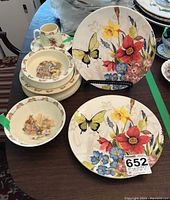Wide view showing three Royal Doulton Bunnykins pieces (two bowls and one cup) and two floral decorative plates with butterfly and flower designs on stands. One Bunnykins plate is also visible, noted to have crack and chip.