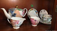 Teapot, cream, sugar bowl, and three teacups with saucers displayed on shelf.