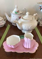 Two white teapots with floral and embossed designs, pink cream and sugar set with tray in foreground on wooden table