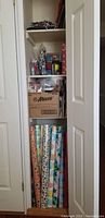 Closet shelves filled with rolls of wrapping paper standing upright, boxes, gift bags, cookie tins, bows, and assorted holiday items visible