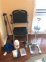 Wide view showing all items arranged on floor and leaning against the wall: resistance chair, cane chair, two canes, neck pillow, white container, spray bottle, grabbing tool, and weighing scale.