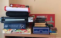 Photo of table and shelf showing mostly boxed board games and card games, including Rummy, Scrabble Junior, multiple decks of cards, and a card shuffler machine. Pavilion Black & White Mexican Train dominoes set also visible.