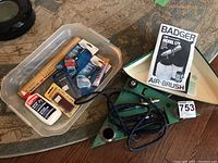 Full view of the model building supplies including the air brush sprayer, various adhesives, brushes, ruler and blades all inside a clear plastic bin on a patterned carpet.