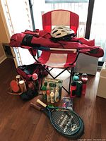 Overall view of the outdoor lot items showing camp chair, helmet, water bottles, insect repellents, tennis rackets, and other small items arranged on a wooden floor near window.