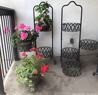 Two black metal plant stands with round lattice-style holders and an extra separate holder, all on a concrete balcony with some plants placed in holders (plants not included in sale).
