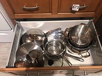Drawer open showing multiple stainless steel pots and pans including lids and a steamer insert. Items are nested and organized inside the drawer.