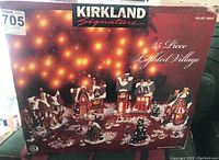 Box front showing 25-piece Kirkland Signature lighted porcelain Christmas village set, model 409810, with festive winter scene and warm glowing lights in each building.