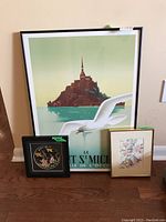 Overall view of three framed art pieces against wall on wood floor, including large Mont Saint Michel print, small floral print and circular silk embroidery.