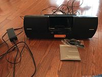 Front view of the black Sirius XM Universal Boombox with power cord and manual placed beside it on a wooden floor.