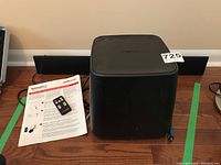 Photo showing the subwoofer, user manual, and remote control on a wooden floor next to the sound bar.