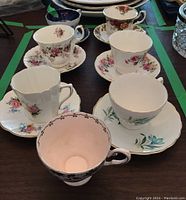 Photo of six teacups with matching saucers and one black and white orphan teacup with floral pattern on wooden table.