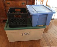 Shows two plastic storage totes stacked on top of each other with a black plastic crate on top. The large beige tote has a green lid labeled 'XMAS'.