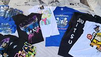 Multiple folded and spread out printed T-shirts in various colors and graphic designs including butterfly, champions, and jiu jitsu prints.