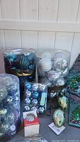Four clear containers of assorted Christmas ornaments in silver, blue, and white colors, one boxed chirping ornament, two hand-decorated egg-shaped glass ornaments, and three green holiday tablecloths visible in packaging.