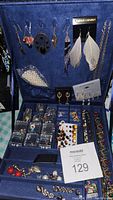 Open blue suede jewelry box showing jewelry hanging on lid, clear plastic cases with earrings, pearl and colorful earrings, bracelets, and multi-color necklaces.