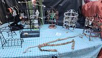 Wide view of eight jewelry displays including black metal stands and two copper-colored earring holders, multiple pieces of jewelry and two wooden bead belts laid out on table covered with blue checkered cloth